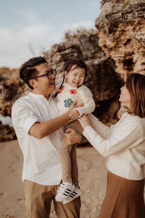 A family in white shirts shares playful moments together on a beach with rocky cliffs in the background.