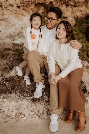 A family sits together on rocky terrain wearing coordinated white and brown outfits in warm sunlight.