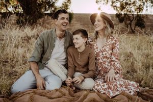 Family of three sitting together on picnic blanket during golden hour in field.