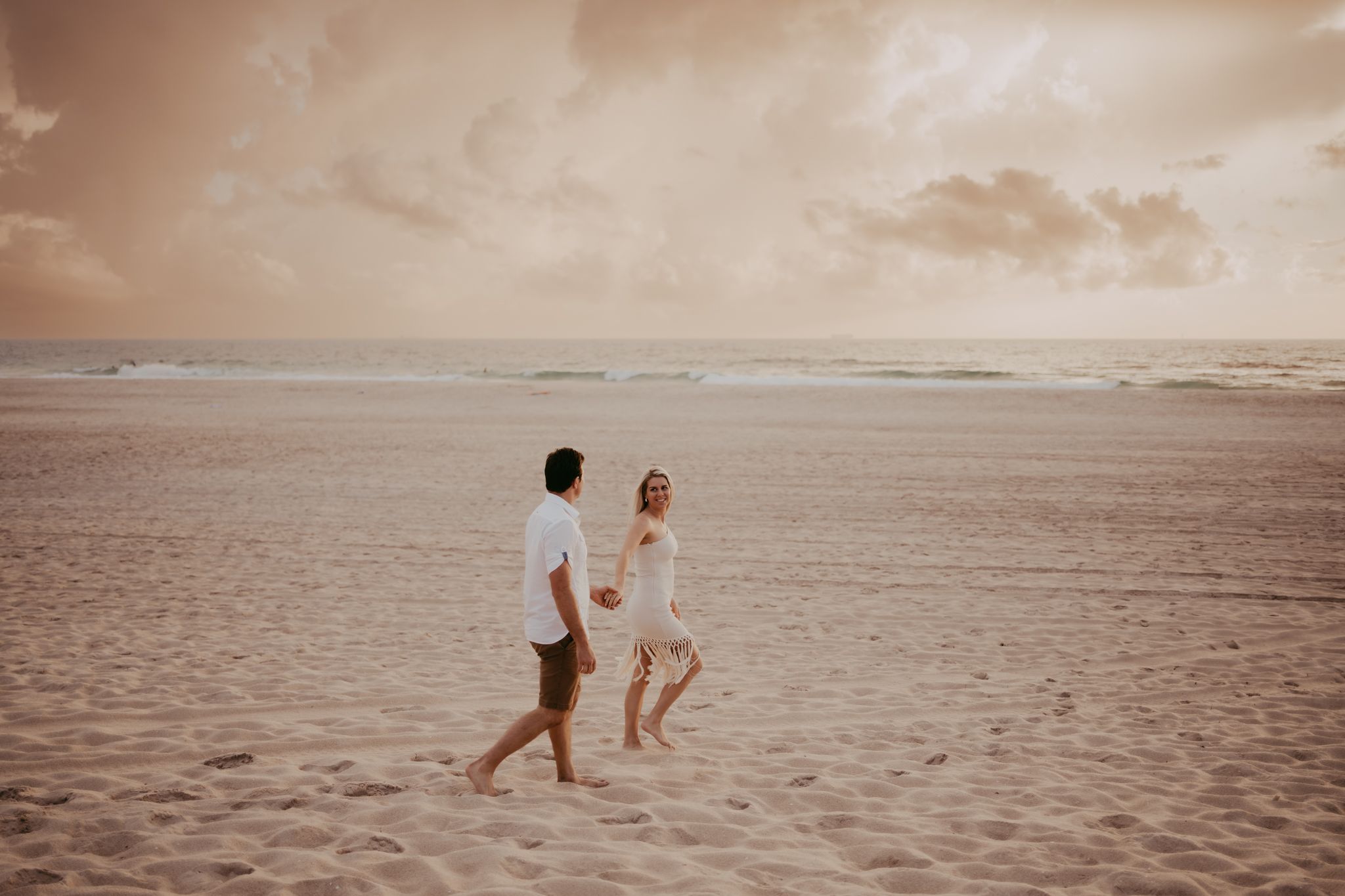 A couple strolls hand in hand on a sandy beach at sunset creating a romantic silhouette against the warm sky.