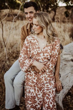 A couple sitting together on a log, with her wearing a floral dress and him in casual brown attire in a golden field.