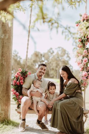 A family sits together on a wooden swing decorated with pink roses in a sun-dappled outdoor setting.