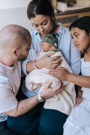 A family gathers tenderly around a sleeping newborn baby swaddled in a soft blanket.