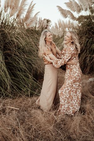 Two women in floral dresses laughing together among tall pampas grass.