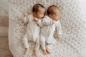 Two babies in white sleepwear lying on a textured cream-colored blanket photographed from above.