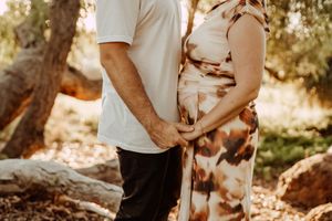 A couple embraces the pregnant belly outdoors in golden sunlight with trees in the background.