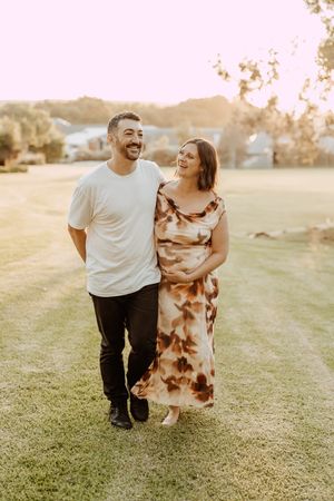 A couple walks together on a grassy field during golden hour sunset, with her wearing a flowing brown patterned dress.