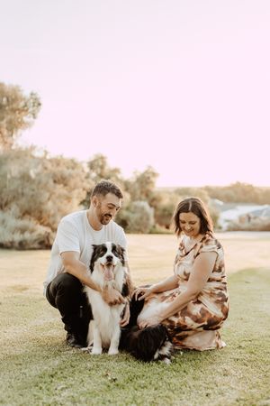 A couple sits on grass with their two dogs during golden hour at sunset in a park setting.