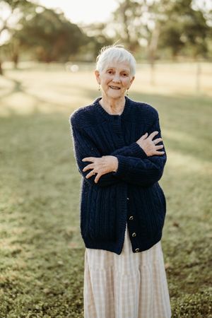 An elderly person in a navy cardigan and striped skirt stands outdoors with crossed arms in soft natural lighting.