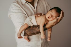 An adult with a tattooed arm gently holds a sleeping newborn in a soft white wrap.