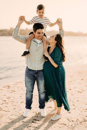 A family playing together on the beach at sunset, with parents supporting their child on shoulders while wearing casual attire.