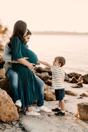 Family maternity photo session on a beach at sunset with rocks and waves creating a serene coastal backdrop.