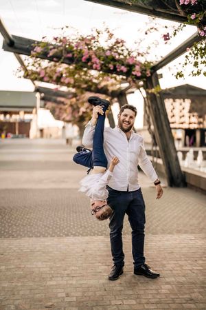 A playful outdoor scene with flowering cherry blossoms overhead and a couple engaged in a fun upside-down pose on a brick plaza.