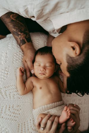 A sleeping newborn in a knit diaper lies on a white textured blanket while being gently held by tattooed arms.