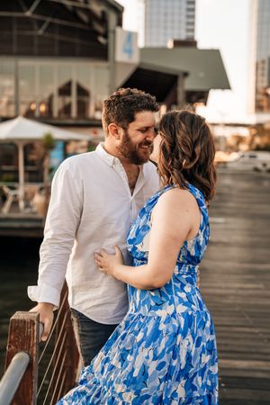 A couple shares an intimate moment on a wooden pier during golden hour, with the woman wearing a blue floral dress.