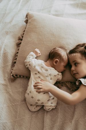 Two children lying on beige bedding share a tender moment as the older one hugs the infant in floral sleepwear.