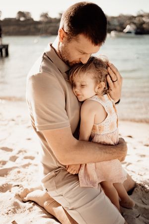 A tender moment between parent and child embracing on a sunny beach with water in the background.