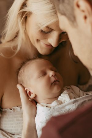 A sleeping newborn baby in a white outfit being lovingly held and admired in a warm, intimate setting.