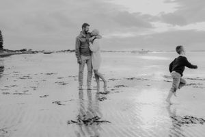 Family plays along beach shoreline in black and white as waves lap at their feet.