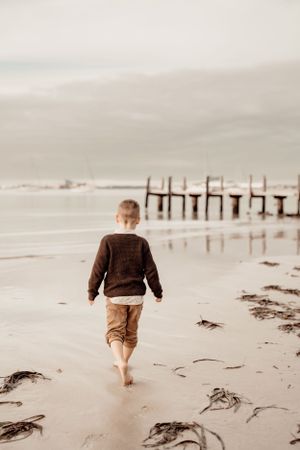 Child walking barefoot on a misty beach toward wooden pier.