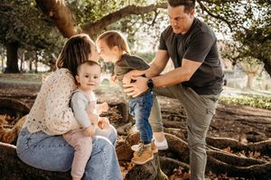A family sits together on tree roots in a sunlit park during a casual outdoor photoshoot.