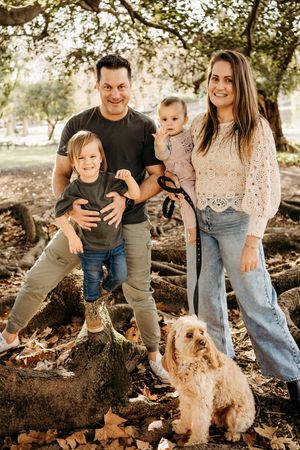 A family poses together outdoors in autumn with their fluffy dog beneath trees with dappled sunlight.