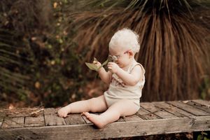 Baby in white onesie sitting on rustic wooden log outdoors surrounded by greenery and natural lighting.