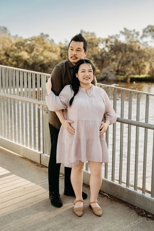 Two people in coordinating pink and black outfits stand together on a wooden deck overlooking trees at sunset.