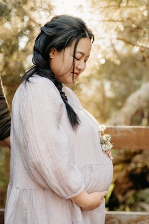 A person in a white dress holds their belly while standing in golden sunlight during a maternity photoshoot.