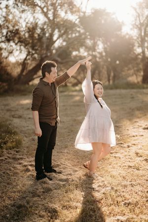 A couple dances playfully together in a golden sunlit field during sunset, creating a romantic outdoor moment.