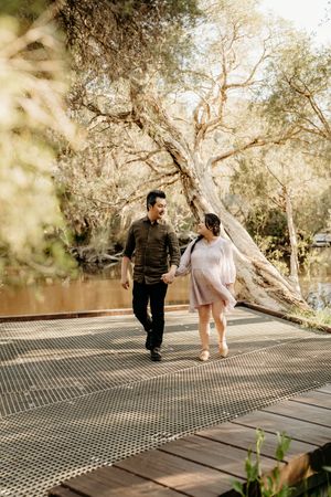 A couple walks together on a wooden boardwalk surrounded by trees in a romantic outdoor setting.