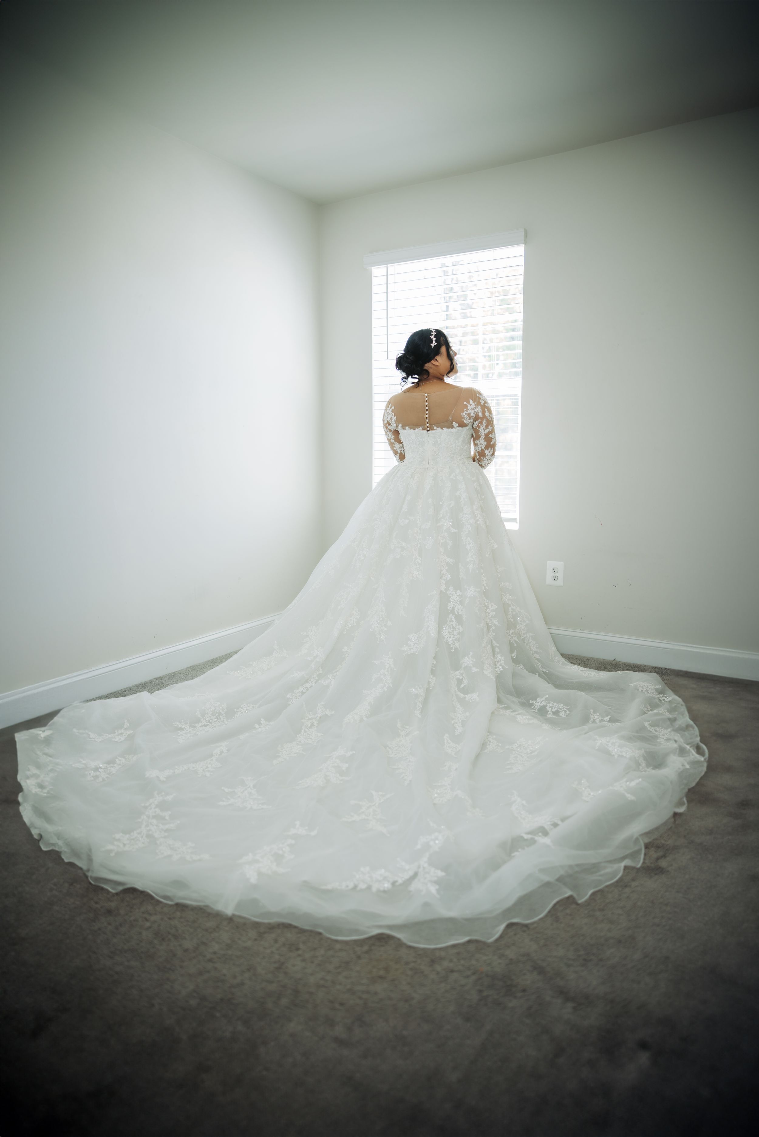 A stunning white wedding dress with lace detailing and long train shown from behind in a minimalist room with natural light.
