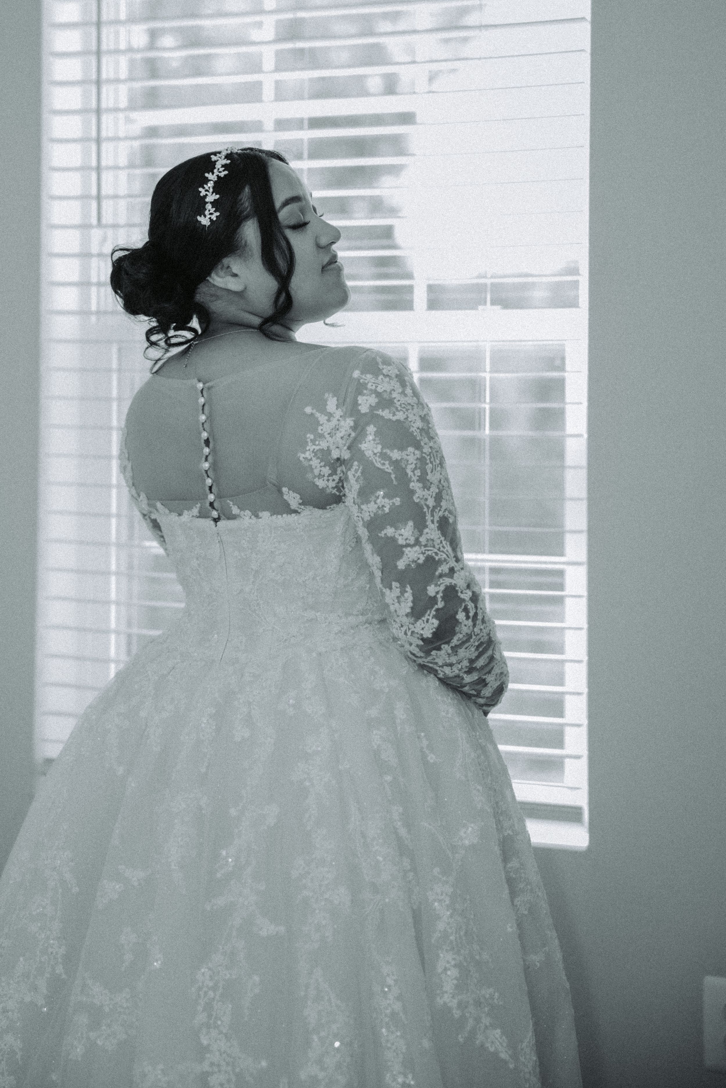 Black and white portrait of a bride in a lace wedding dress with long sleeves looking out a window by venetian blinds.