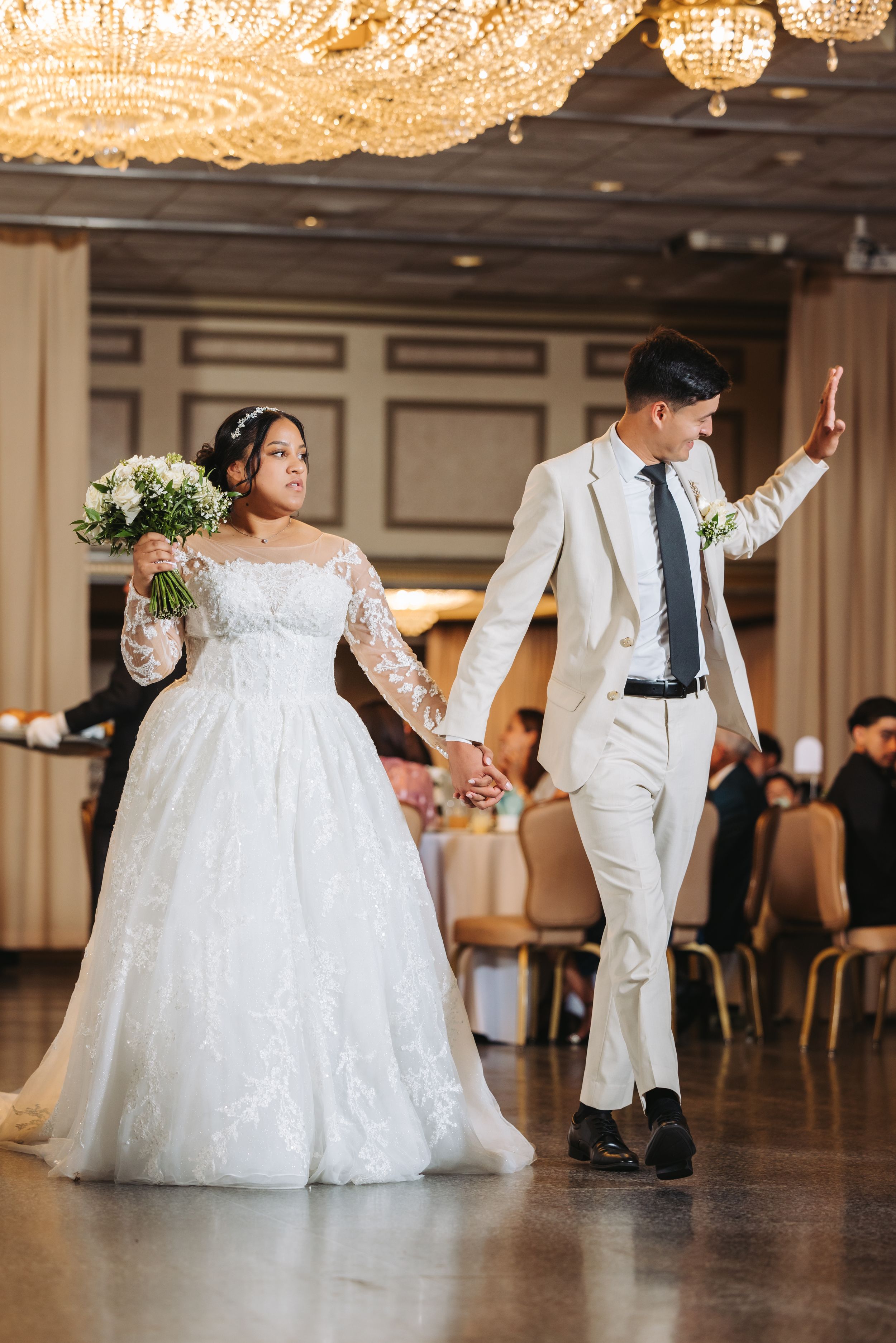 Newlyweds share a joyful moment walking hand in hand at their wedding reception under crystal chandeliers.