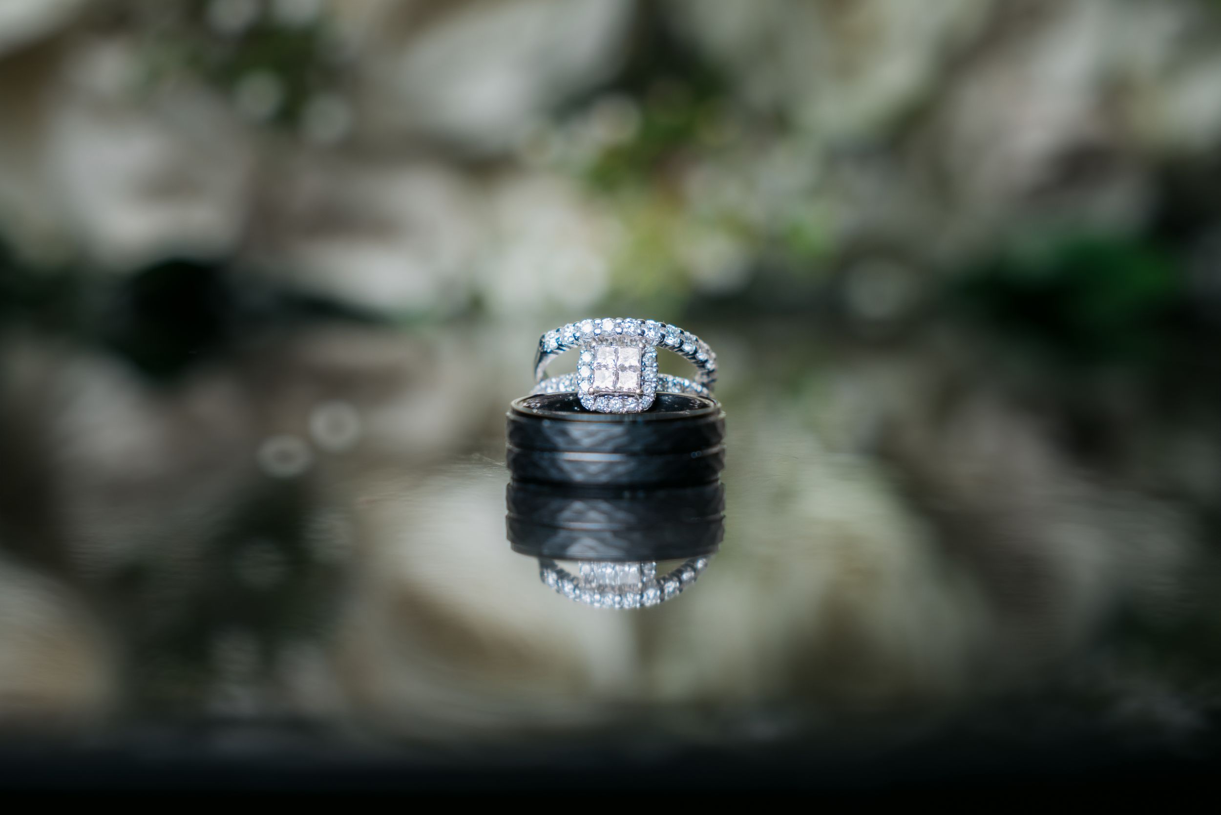 A sparkling diamond engagement ring is reflected on a dark mirrored surface with a blurred natural background.