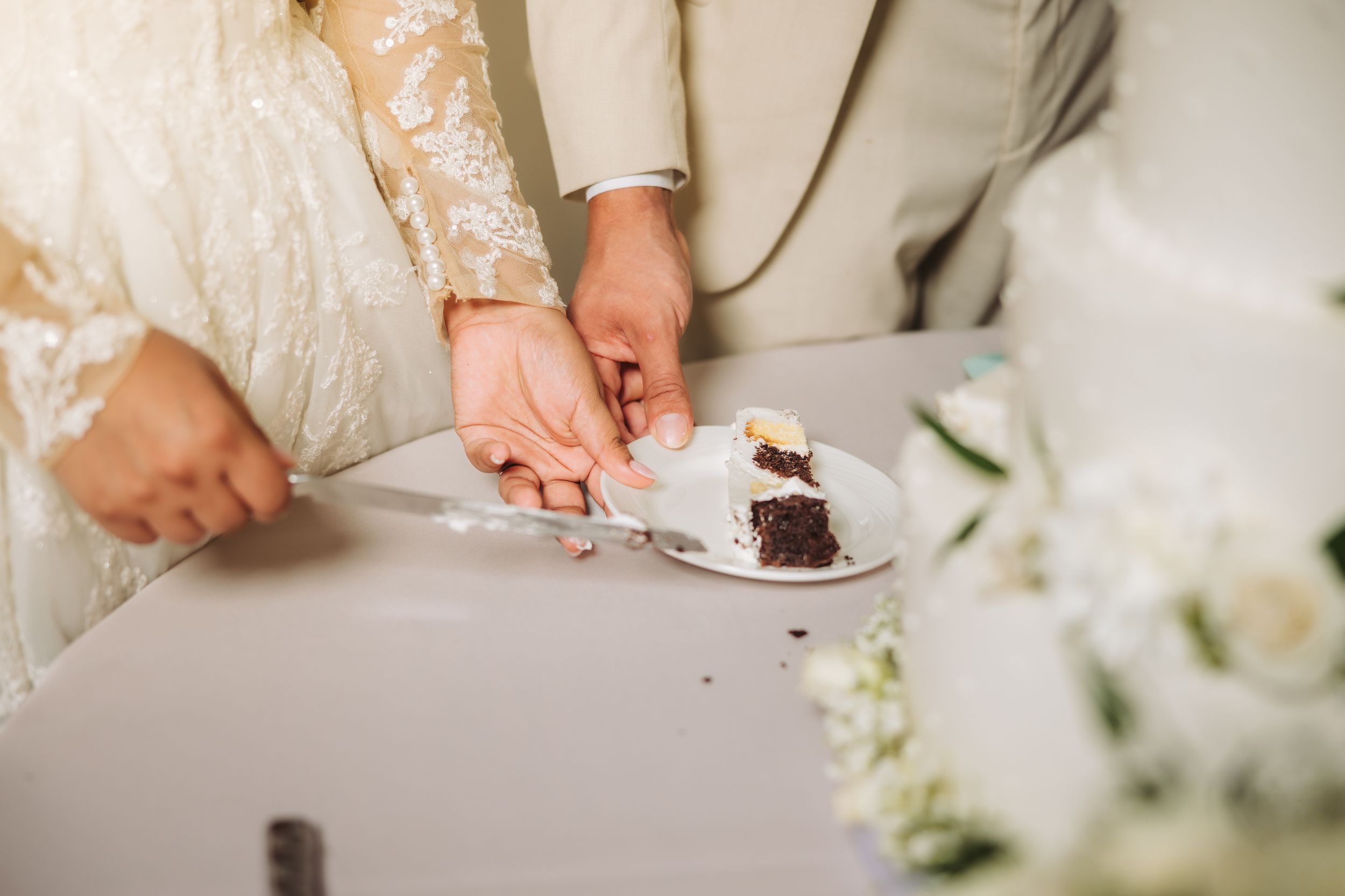Hands cutting into a slice of chocolate wedding cake surrounded by white floral decorations on a light tablecloth.