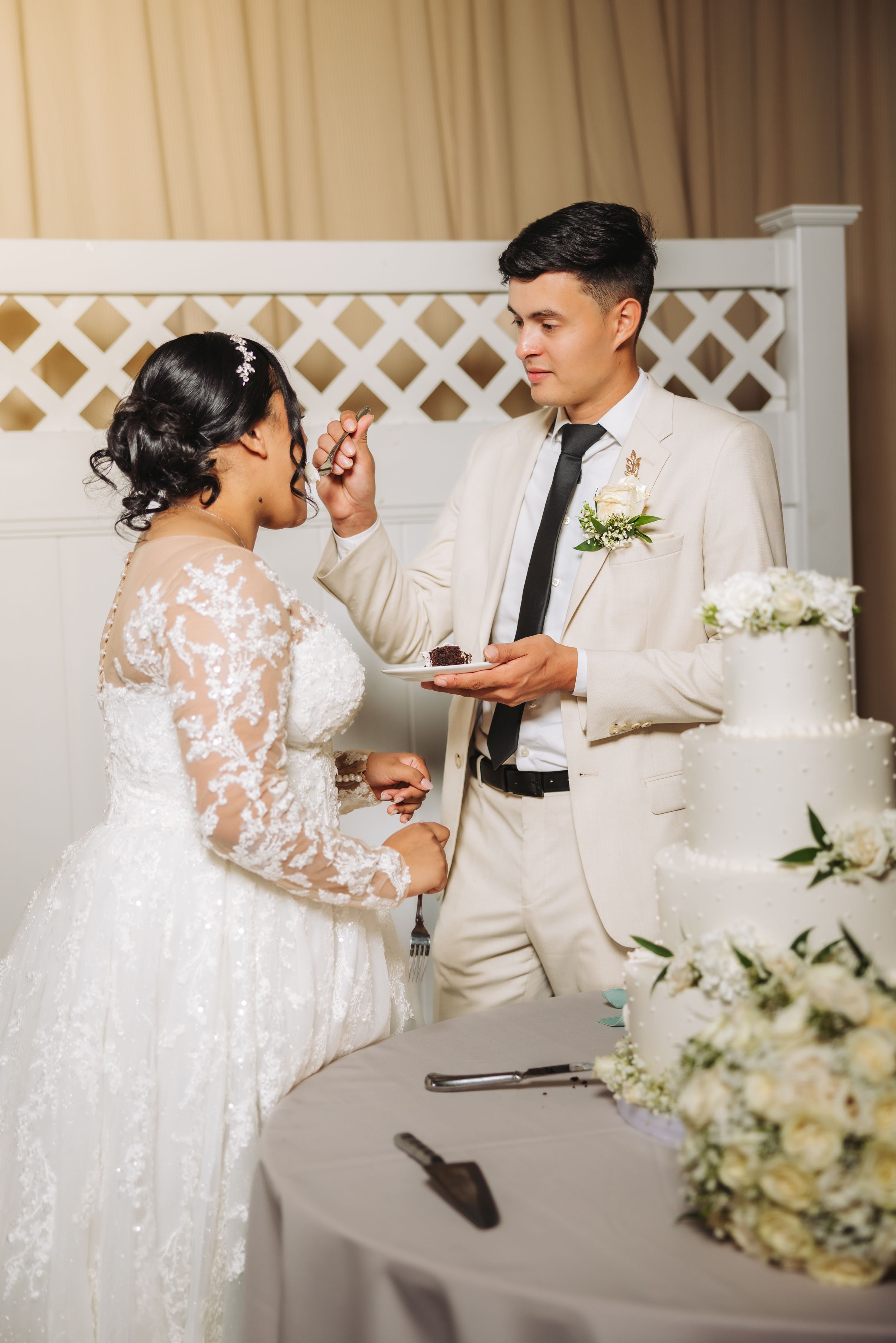 A couple in elegant wedding attire share a piece of white tiered wedding cake at their reception ceremony.