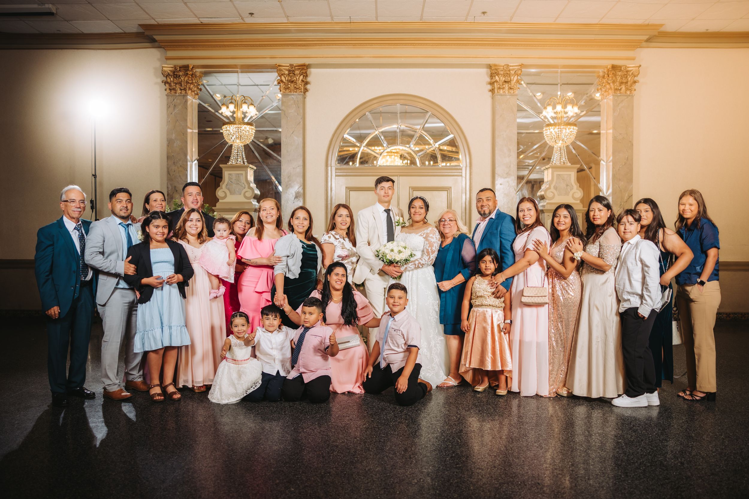 A large group poses together in formal attire at an elegant venue with ornate gold columns and arched details.