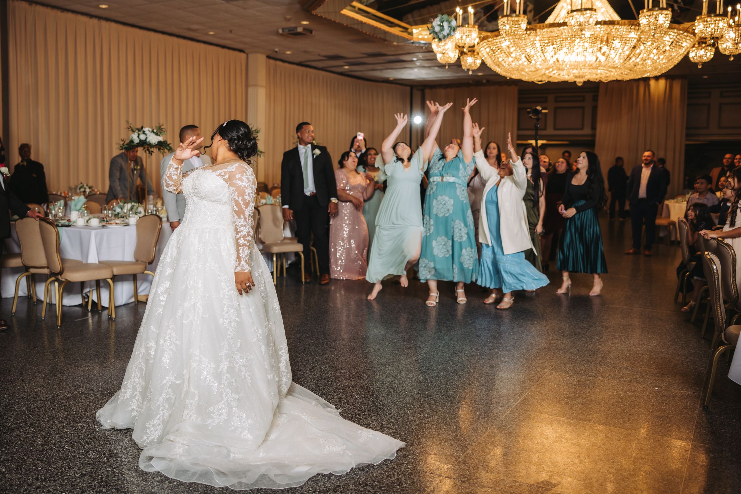 A wedding reception scene shows guests in blue dresses reaching for a bouquet toss while others look on in an elegant ballroom.