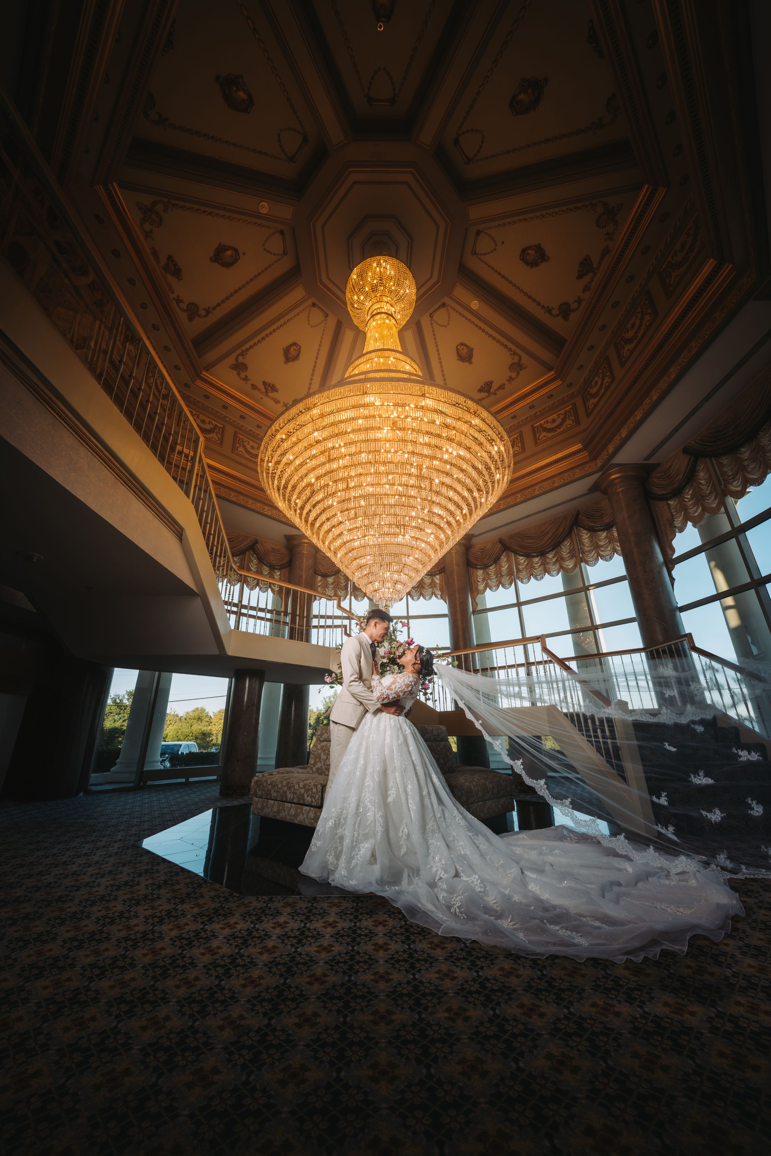 Elegant ballroom with ornate crystal chandelier and patterned ceiling features a couple embracing beneath the golden light.