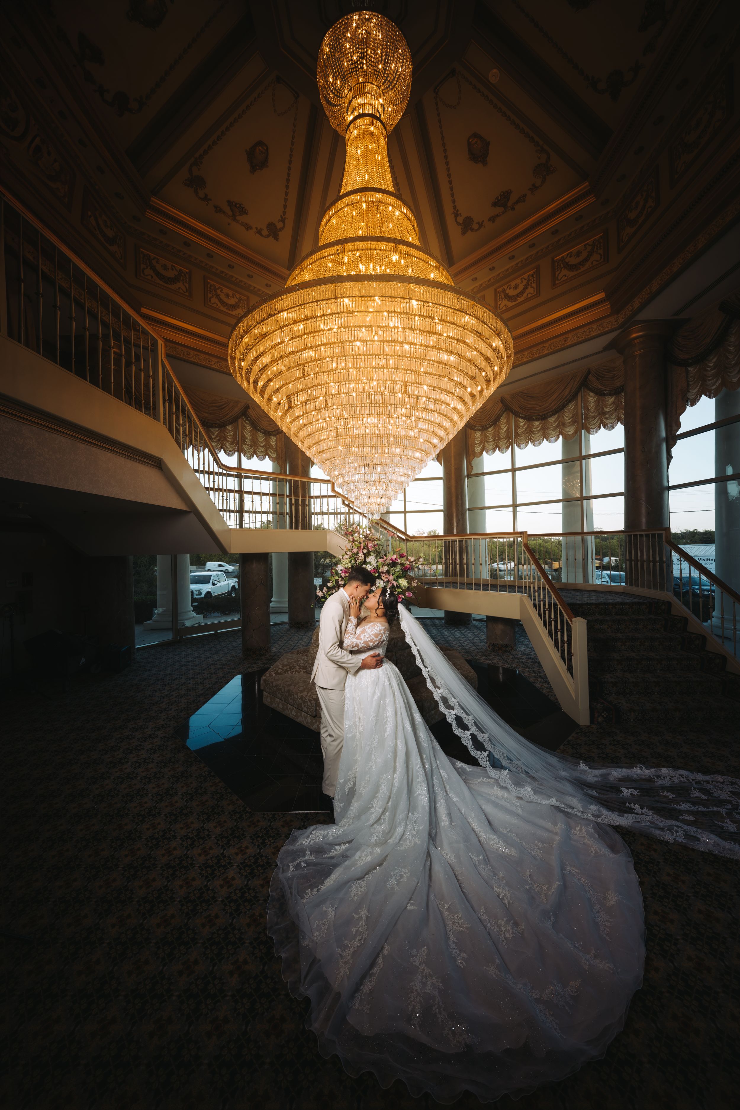 Elegant wedding scene featuring a flowing white gown and long veil under a grand crystal chandelier in an ornate ballroom.