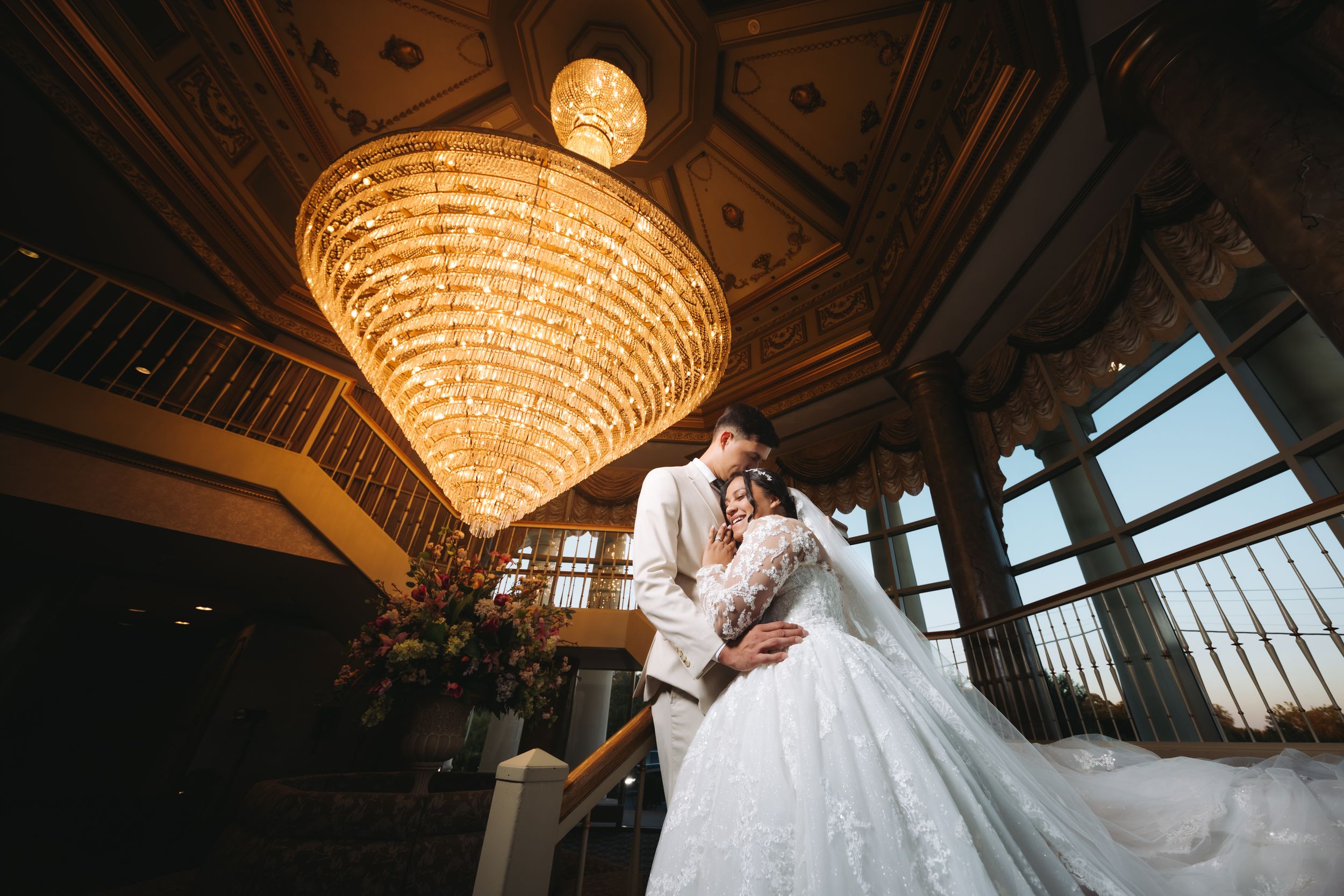 A couple shares an intimate moment beneath an ornate crystal chandelier in an elegant ballroom with large windows.