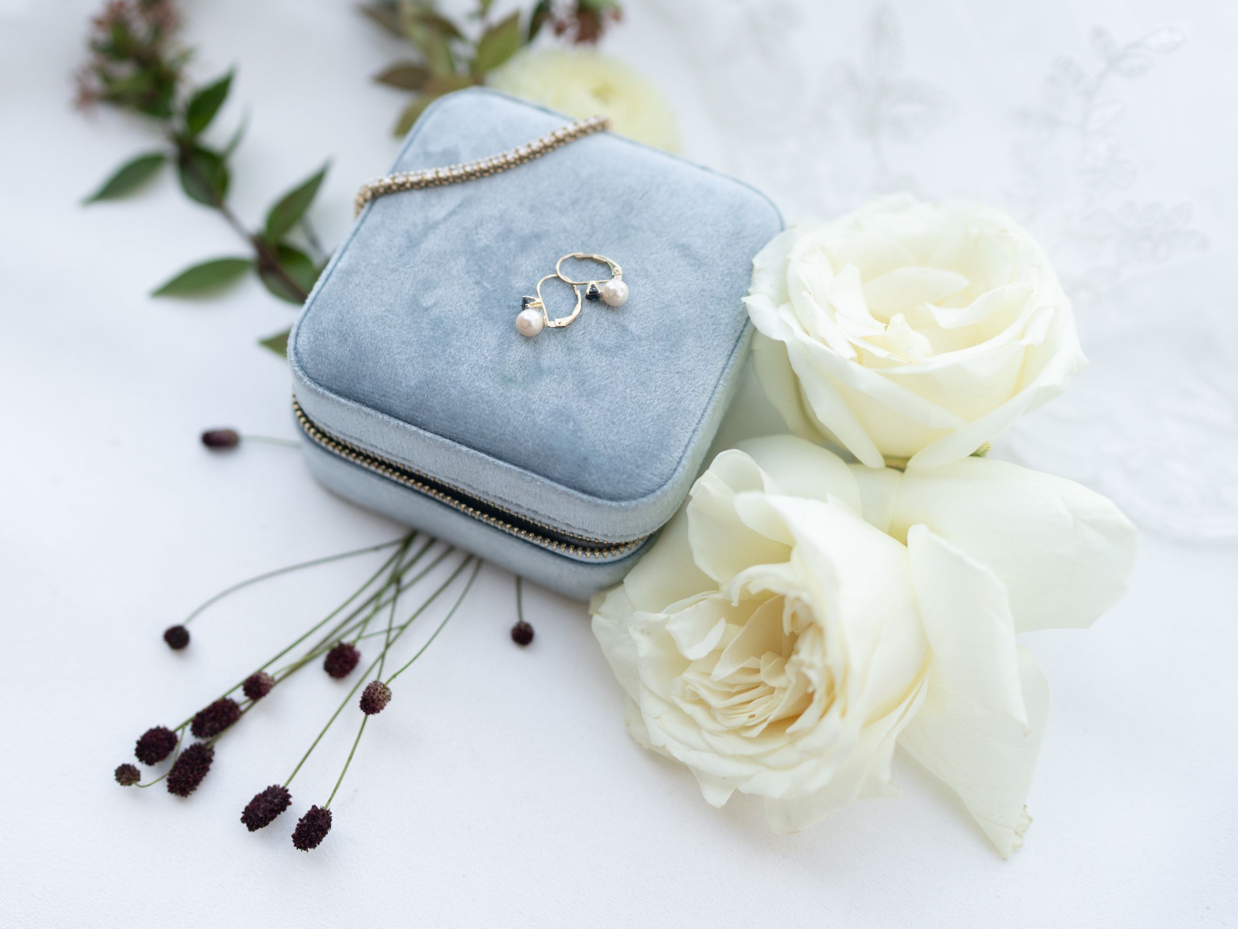 Silver earrings rest in a light blue velvet box beside delicate white roses and decorative brown stems.