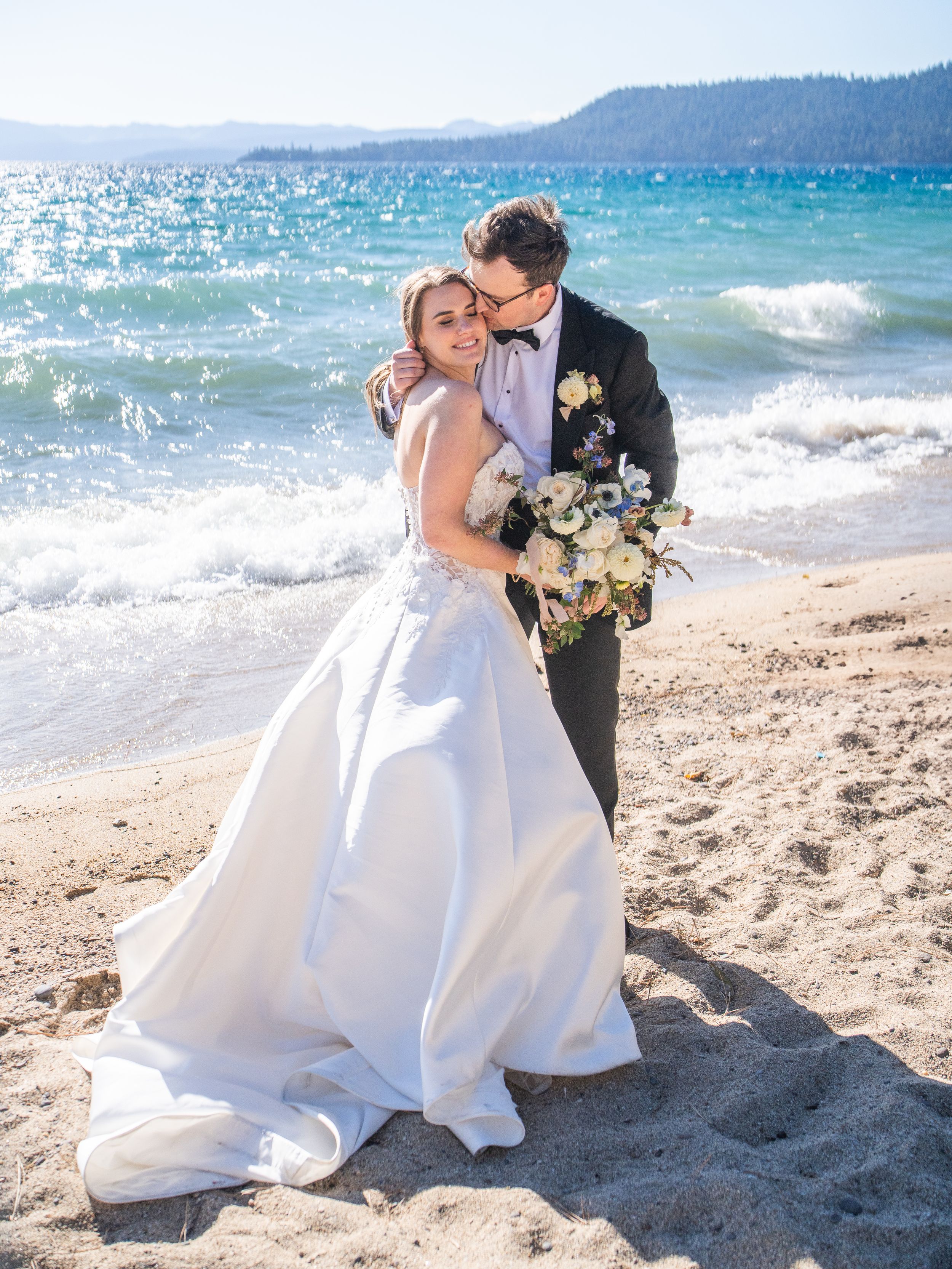 A couple in wedding attire embrace on a sunny beach at Hyatt Lake Tahoe with sparkling turquoise water and mountains in the distance.