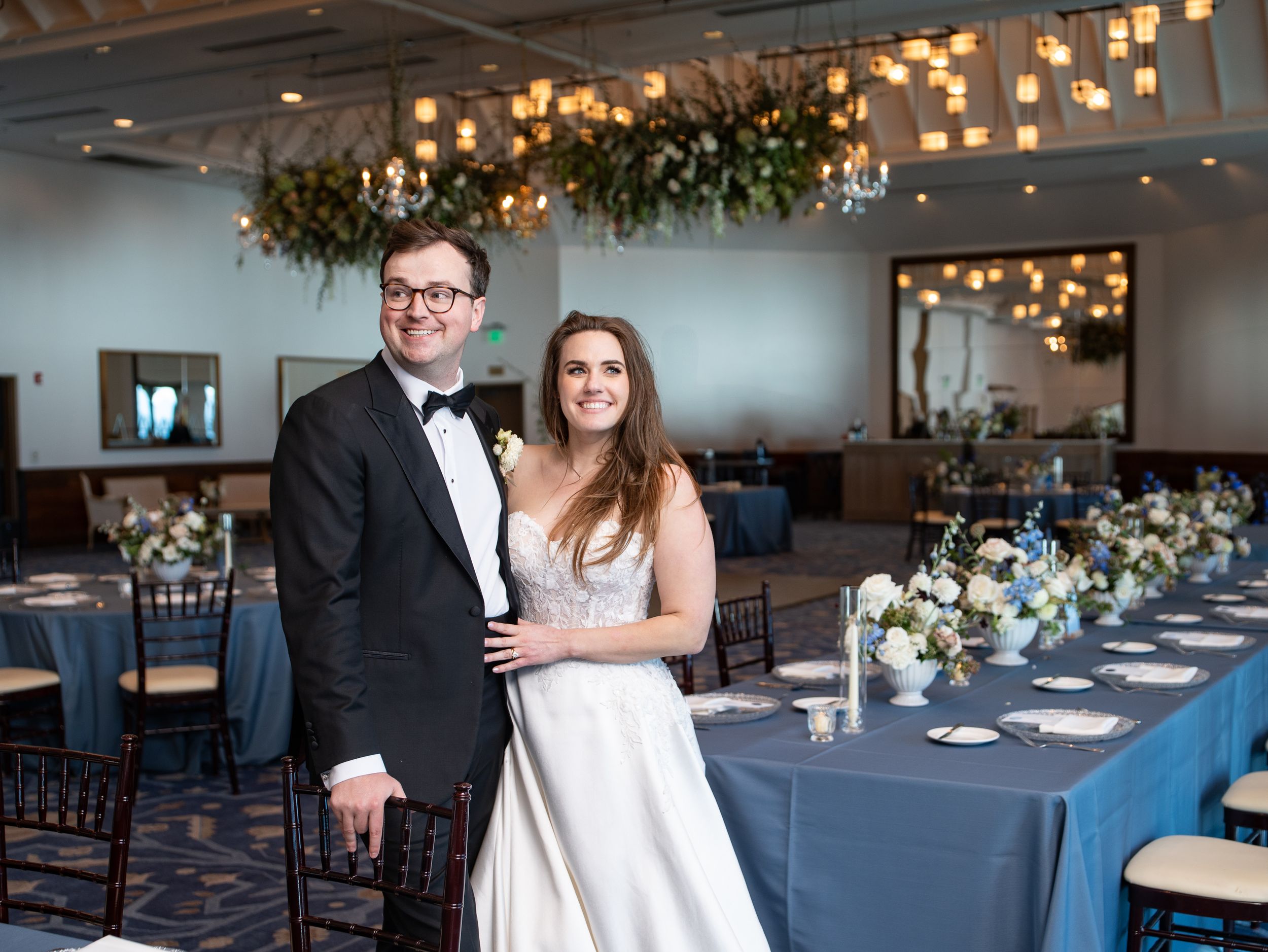 A happy couple poses in formal wedding attire inside an elegant lakeside ballroom at Hyatt with blue table settings and overhead lighting.
