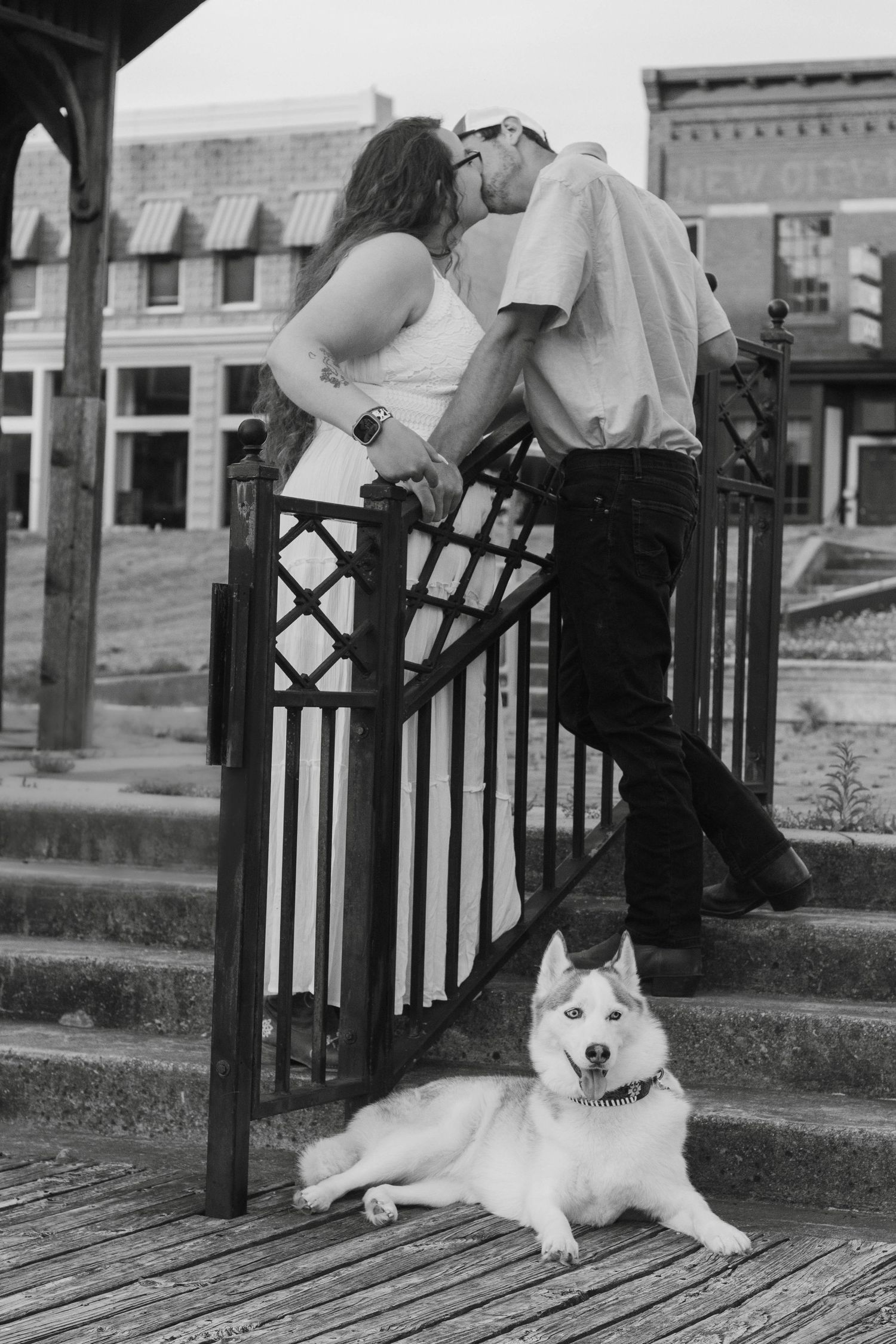 A couple shares a romantic moment on steps with a white dog sitting below in black and white.