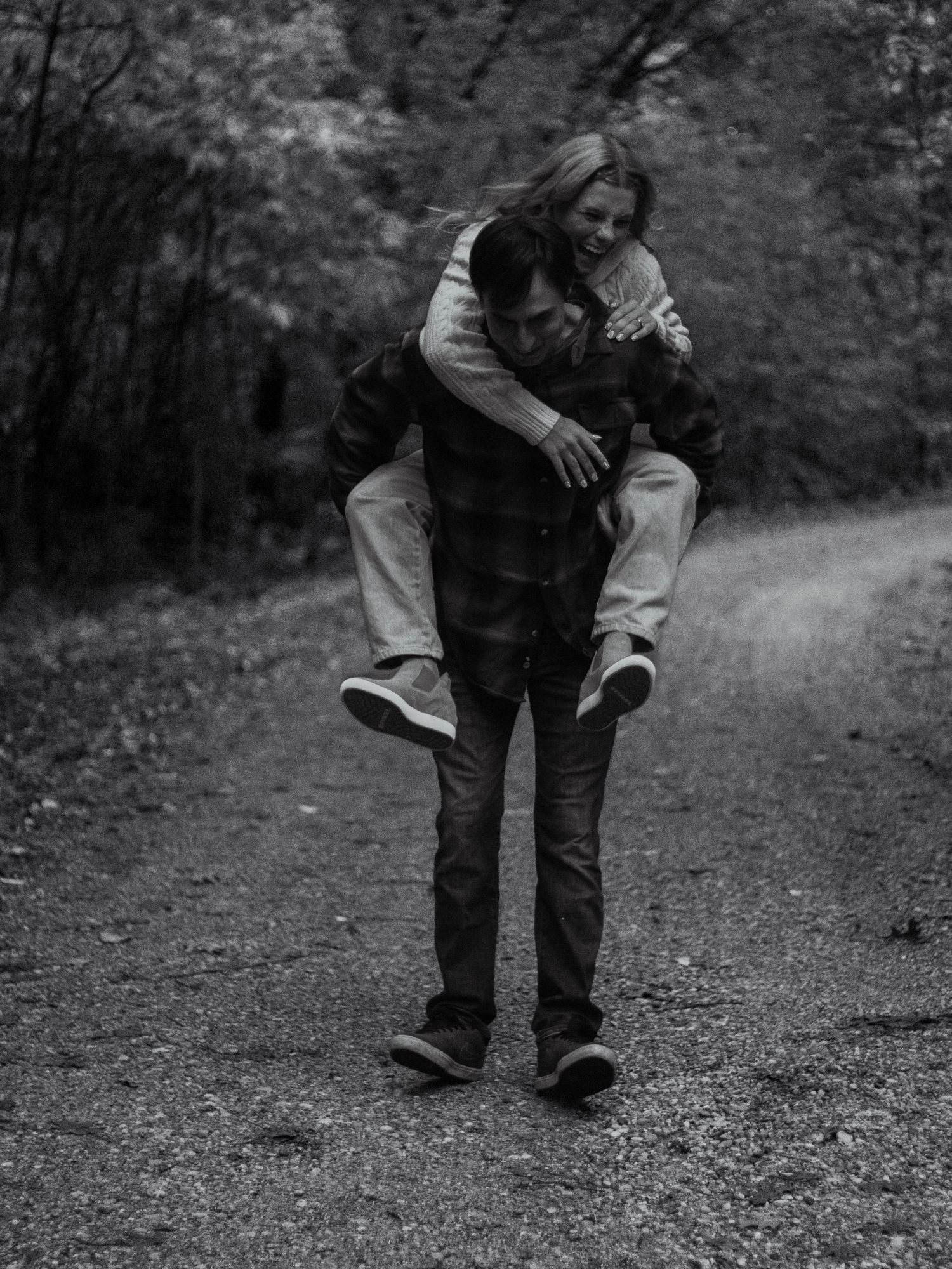 A couple shares a piggyback ride moment on a wooded trail captured in black and white photography.