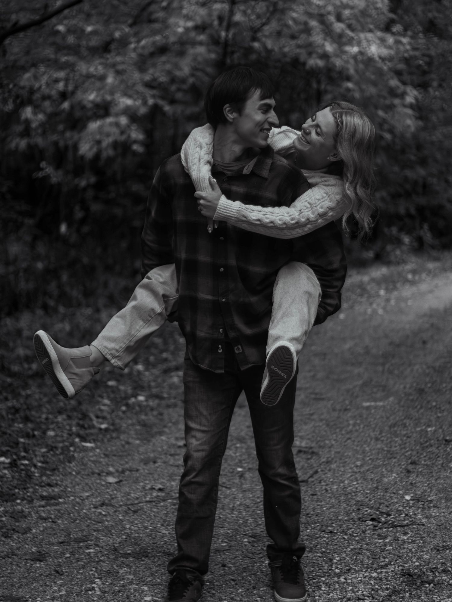 A romantic moment captured in black and white as a couple plays and laughs together on a forest path.