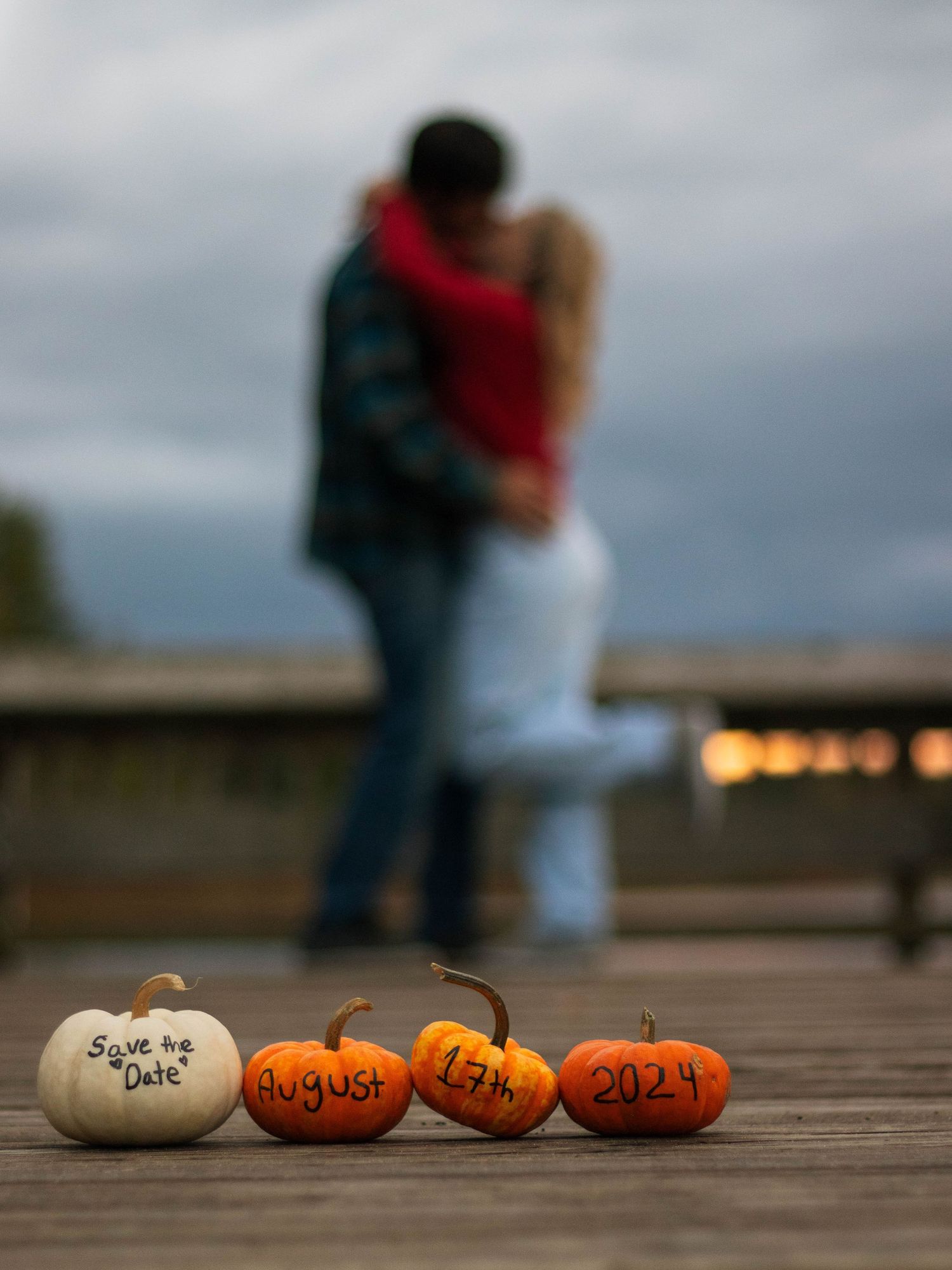 Couple silhouetted against dusk sky with 'Save the Date' pumpkins displayed on wooden dock.