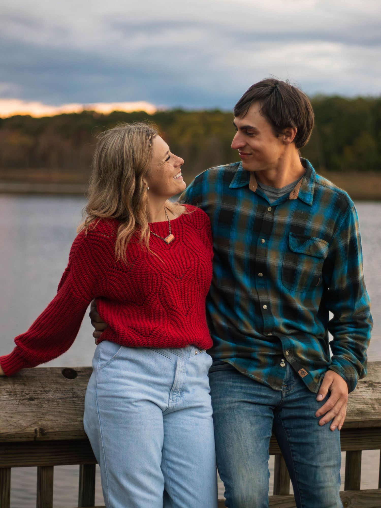 A couple shares a loving moment on a dock with a red sweater and plaid shirt against a moody sky.